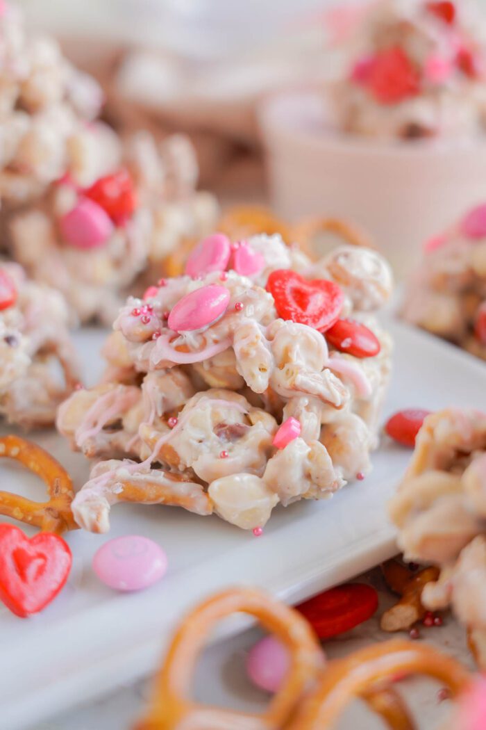 A close-up of a white chocolate cluster with pretzels, pink and red candies, and heart-shaped sprinkles on a white plate with additional clusters and pretzels in the background.