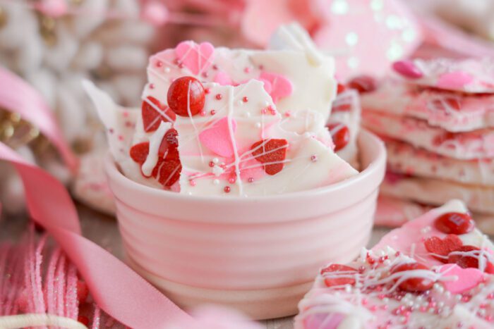 A bowl of white chocolate bark pieces adorned with pink and red candies, sprinkles, and drizzle, surrounded by additional bark pieces and pink ribbons.