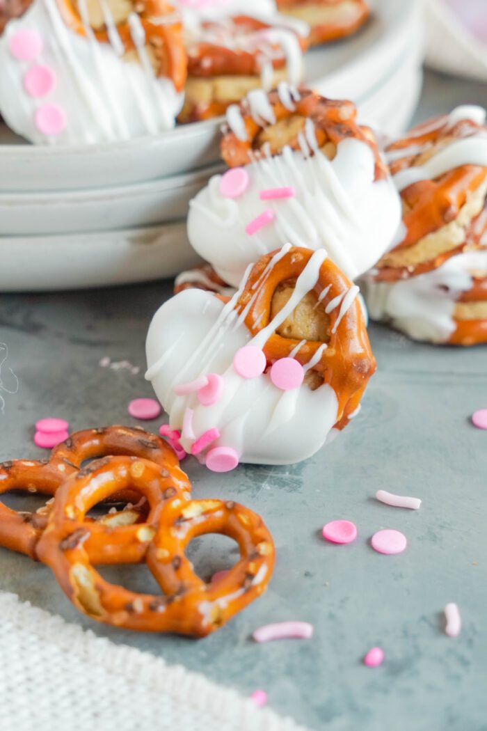 A close-up of pretzels dipped in white chocolate and decorated with pink sprinkles, arranged on a gray surface. Additional undipped pretzels and pink sprinkles are scattered around.