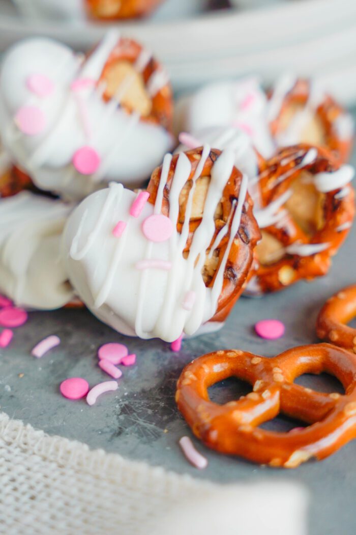 White chocolate-covered pretzels with pink sprinkles are piled on a gray surface, surrounded by additional whole pretzels and pink decorative sprinkles.