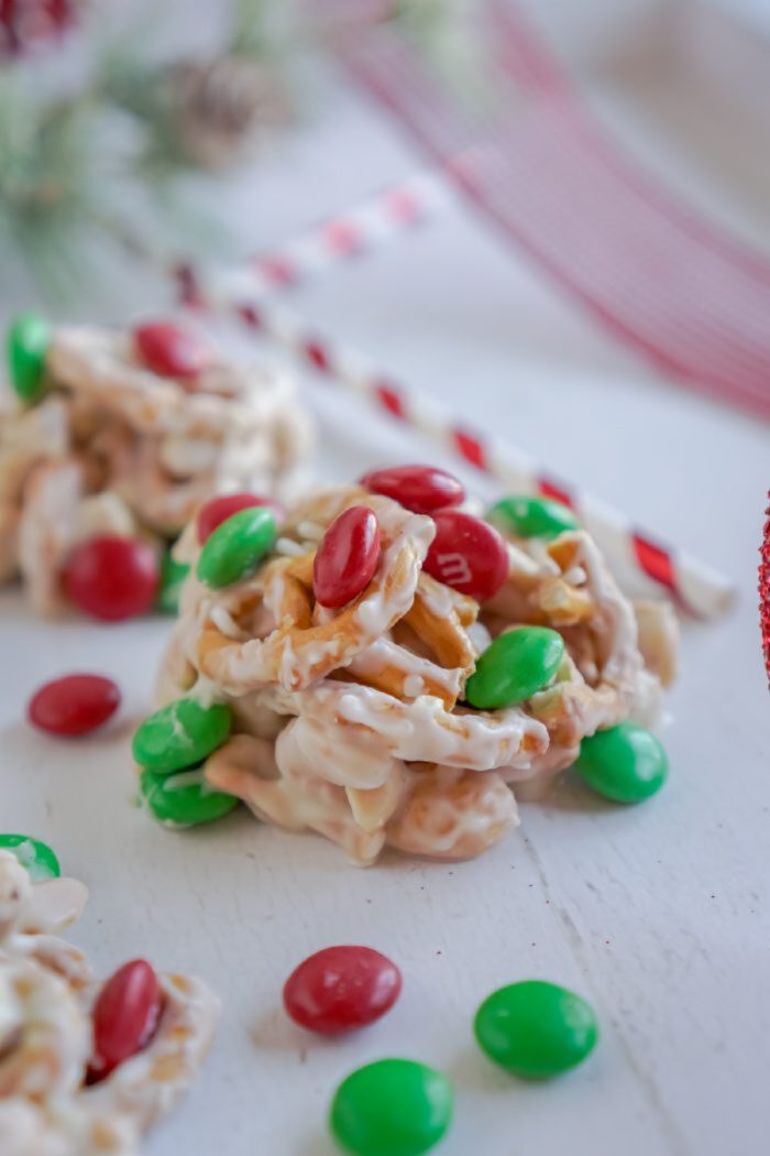 A cluster of white chocolate-coated cereals and nuts with red and green candies on top sits on a white surface, surrounded by a few scattered candies.