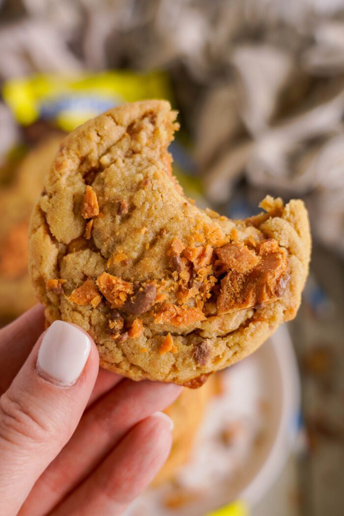 A hand holding a partially eaten cookie with visible chunks of butterfinger on a blurred background.