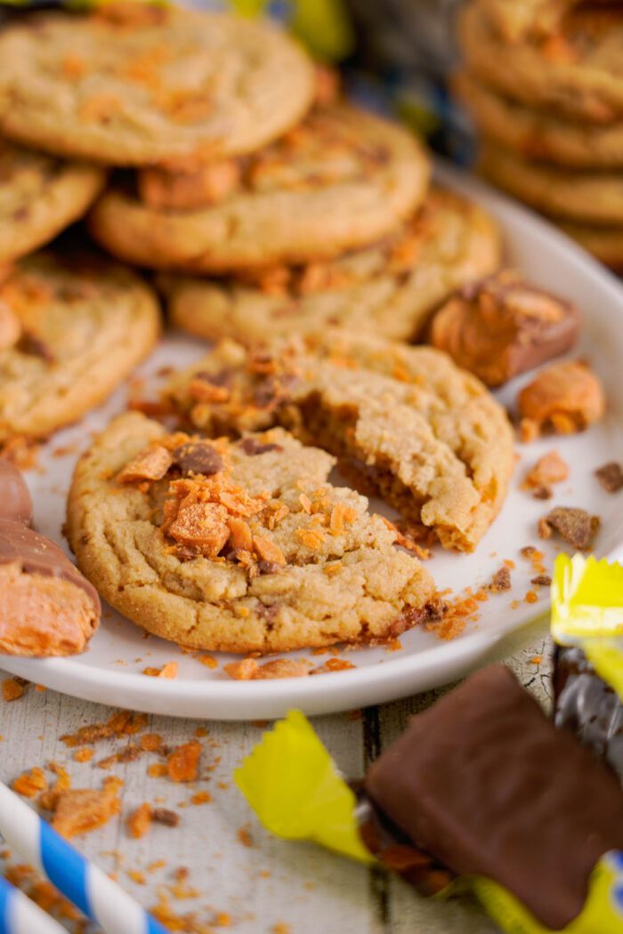 A white plate holds several cookies with scattered chunks of butterfinger pieces on them. One cookie is broken in half. Unwrapped butterfingers are placed beside the plate.