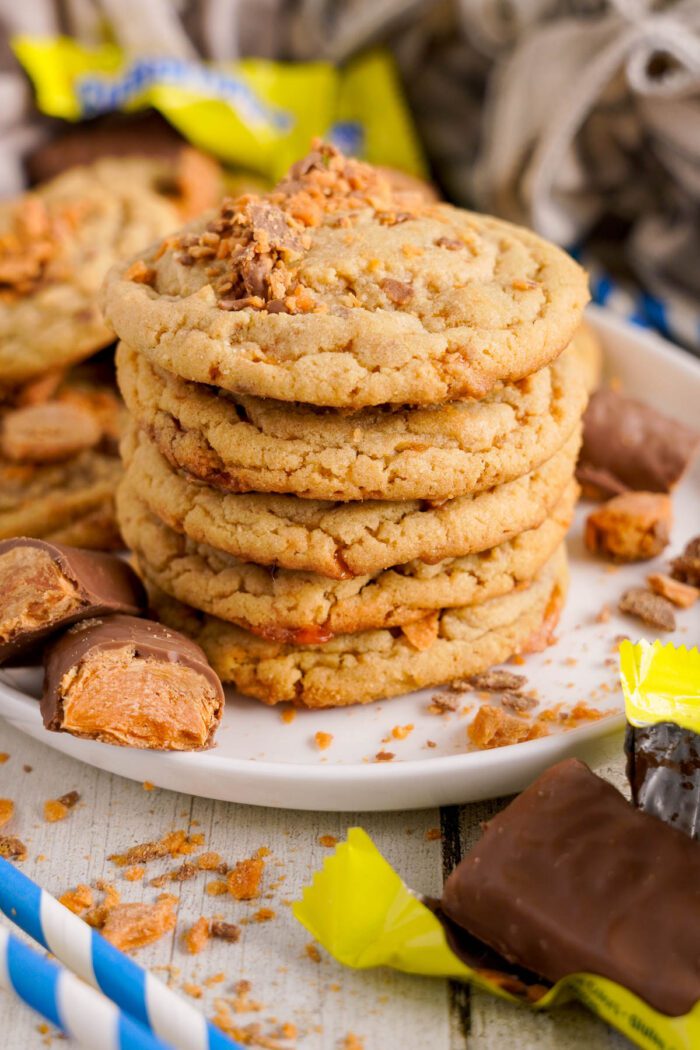 A stack of cookies with crumbled candy pieces on a white plate, surrounded by unwrapped chocolate-covered candies and yellow wrappers on a wooden surface.