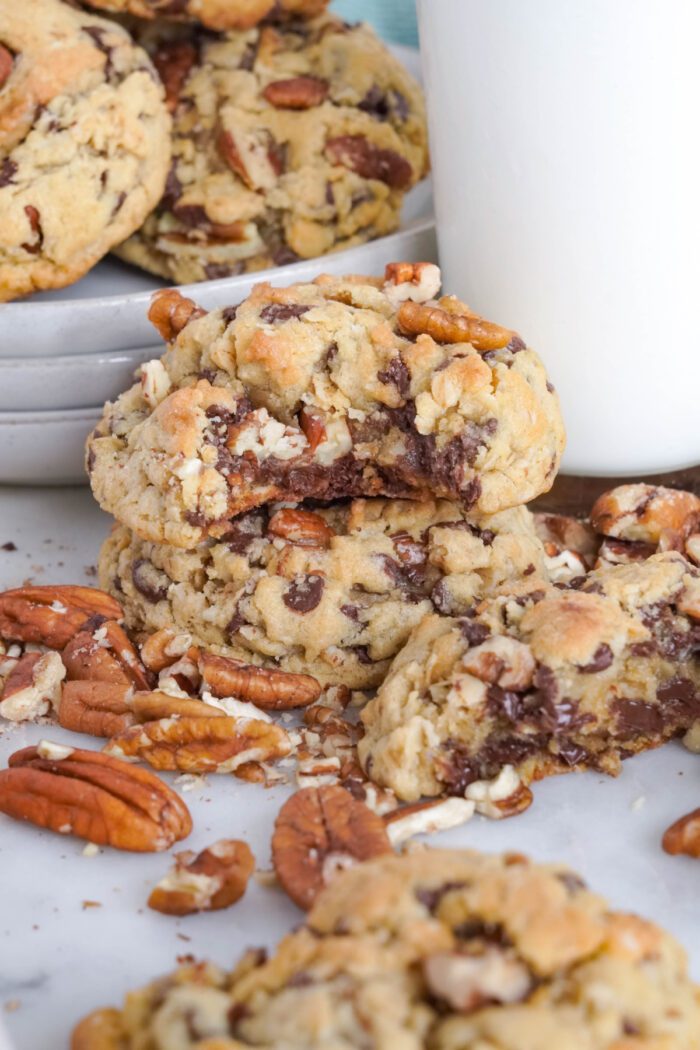 Close-up of pecan chocolate chip cookies on a white surface, with a pile of cookies in the background. Some pecan halves and crumbs are scattered around, and a glass of milk is partially visible.