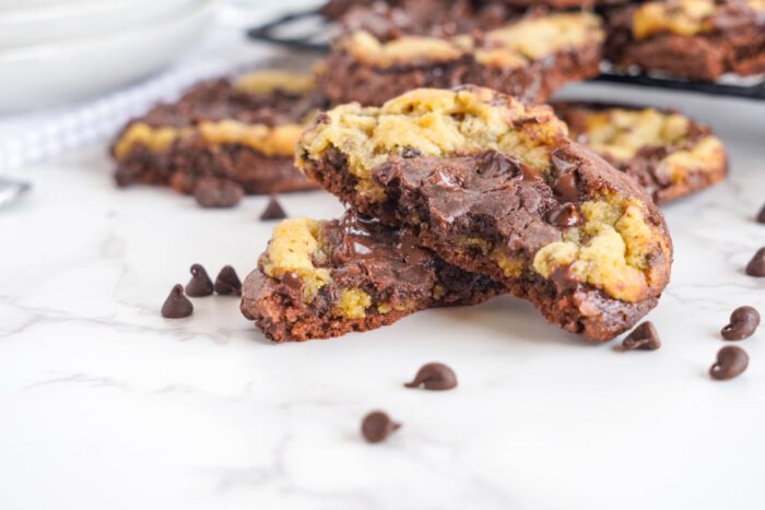 A close-up of a chocolate marbled cookie split in half, revealing melted chocolate chips inside. More cookies and scattered chocolate chips are visible in the background on a marble surface.