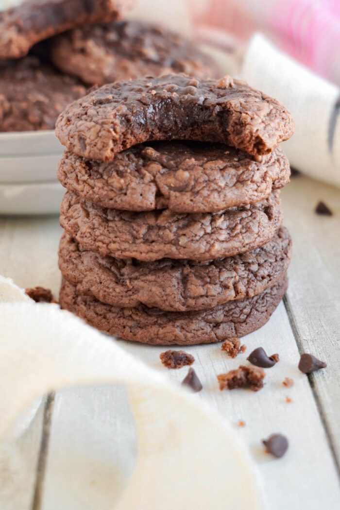 Stack of four chocolate cookies on a wooden surface, with the top cookie partially eaten and chocolate chips scattered around.