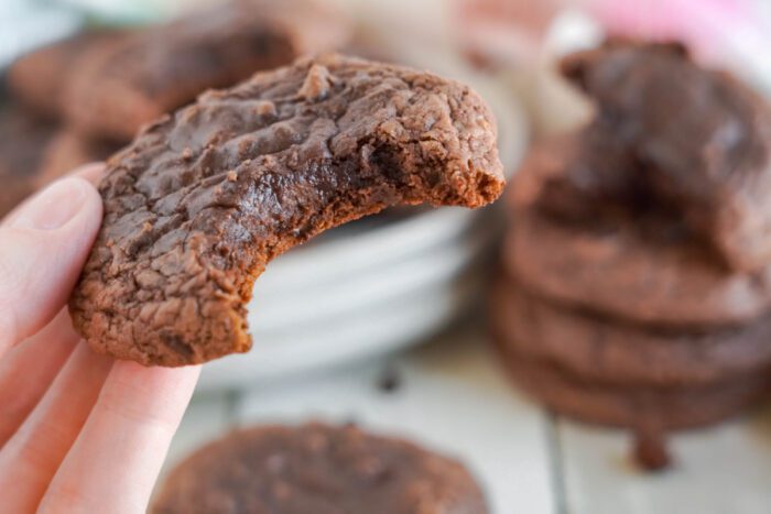 A hand holds a chocolate cookie with a bite taken out of it. Blurry cookies are stacked in the background.