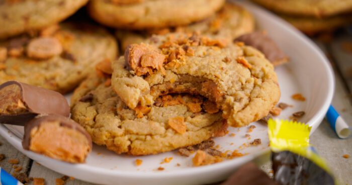 Close-up of a plate holding butterfinger cookies, one of which has a bite taken out of it. Crumbled pieces of butterfinger and crumbs are scattered around.