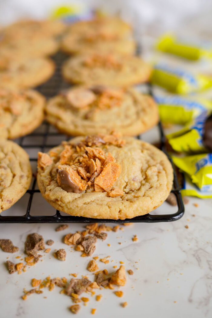 A cooling rack with cookies topped with crushed butterfinger pieces. Some crumbs are scattered on the marble countertop. In the background are yellow butterfinger candy wrappers.