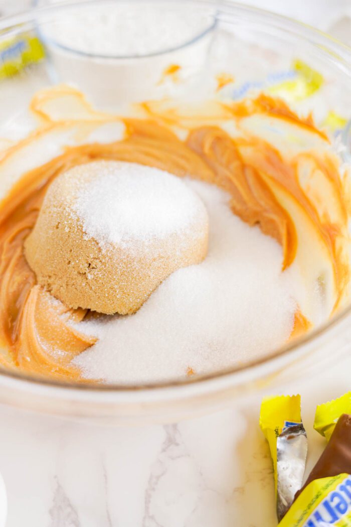 A mixing bowl containing creamy peanut butter, brown sugar, and white sugar, with a cup of flour and candy wrappers in the background on a marble countertop.