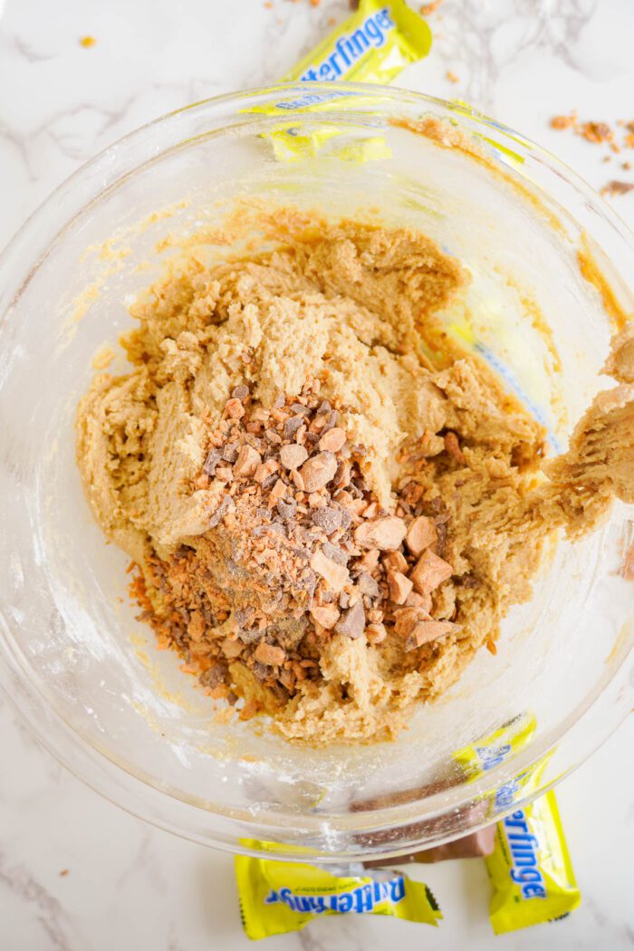 A clear bowl with cookie dough mixed with crushed Butterfinger candy pieces on a white marble surface. Two Butterfinger candy wrappers are partially visible around the bowl.