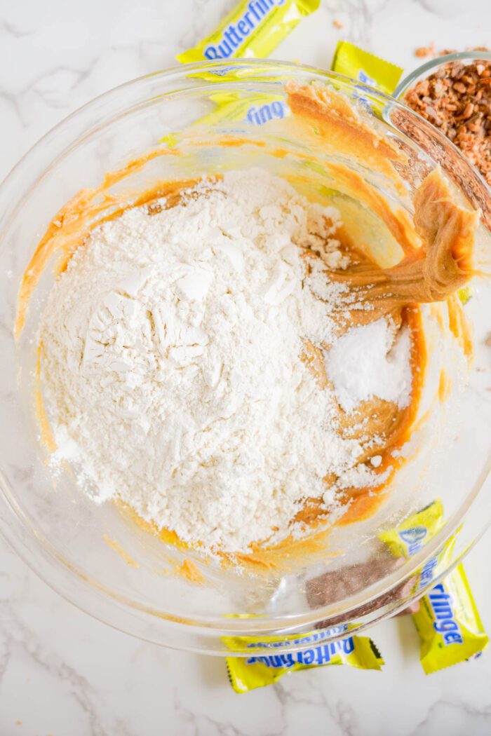 A mixing bowl containing baking ingredients like flour and peanut butter. Wrappers of Butterfinger candy and chopped Butterfinger pieces are nearby.