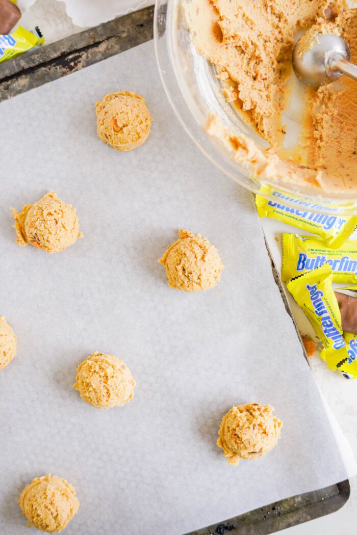 Scoops of cookie dough on parchment paper are displayed, with a mixing bowl of dough and several candy wrappers nearby.