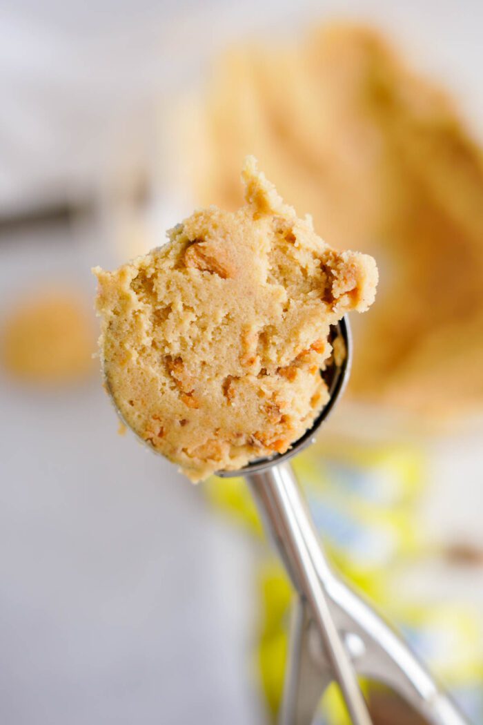 Close-up of a metal ice cream scoop containing raw cookie dough with small chunks, blurred background showing a dough ball and rolling pin.