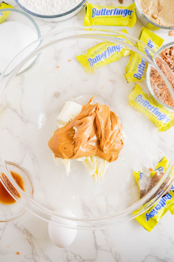 A glass bowl contains peanut butter and cream cheese, surrounded by Butterfinger candy wrappers, vanilla extract, an egg, and other baking ingredients on a marble countertop.