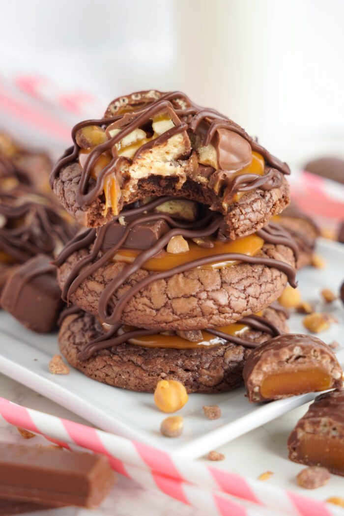 A stack of chocolate cookies with caramel and chocolate drizzle, topped with nuts. Some cookies are partially eaten, and there are additional cookies and striped straws in the background.