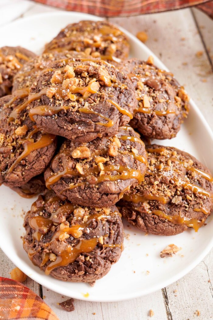 A plate of chocolate cookies topped with caramel drizzle and crushed nuts, placed on a white surface.