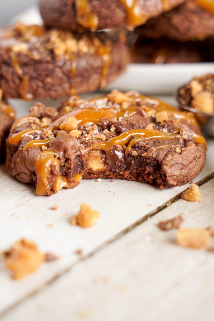 A close-up of a partially eaten chocolate cookie drizzled with caramel and sprinkled with nuts, with crumbs scattered on a wooden surface. Other cookies are visible in the background, perfect inspiration for those seeking recipes with chocolate chips.