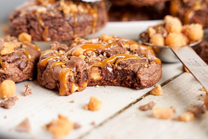 A close-up of a chocolate cookie with caramel drizzle and nuts, partially eaten. Additional uncrushed nuts are in a spoon nearby.