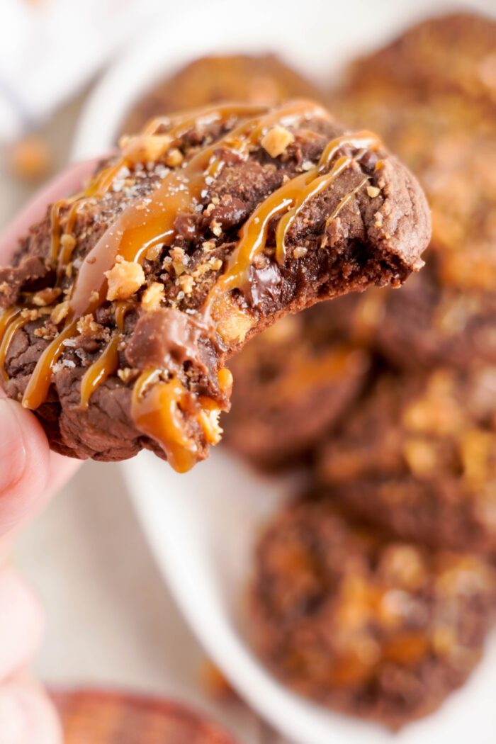 A close-up of a hand holding a chocolate cookie with caramel drizzle and nuts, showing a bite taken out. More cookies are blurred in the background on a white plate.