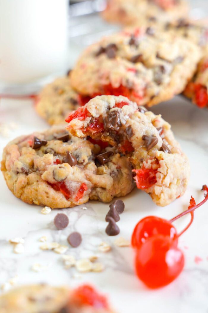 Close-up of chocolate chip cookies with visible pieces of red cherries and oats on a white surface. A glass of milk is partially visible in the background. Some chocolate chips and oats are scattered around.