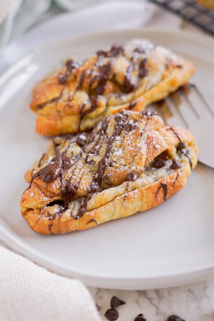Two chocolate-drizzled pastries are placed on a white ceramic plate. The pastries are sprinkled with powdered sugar.