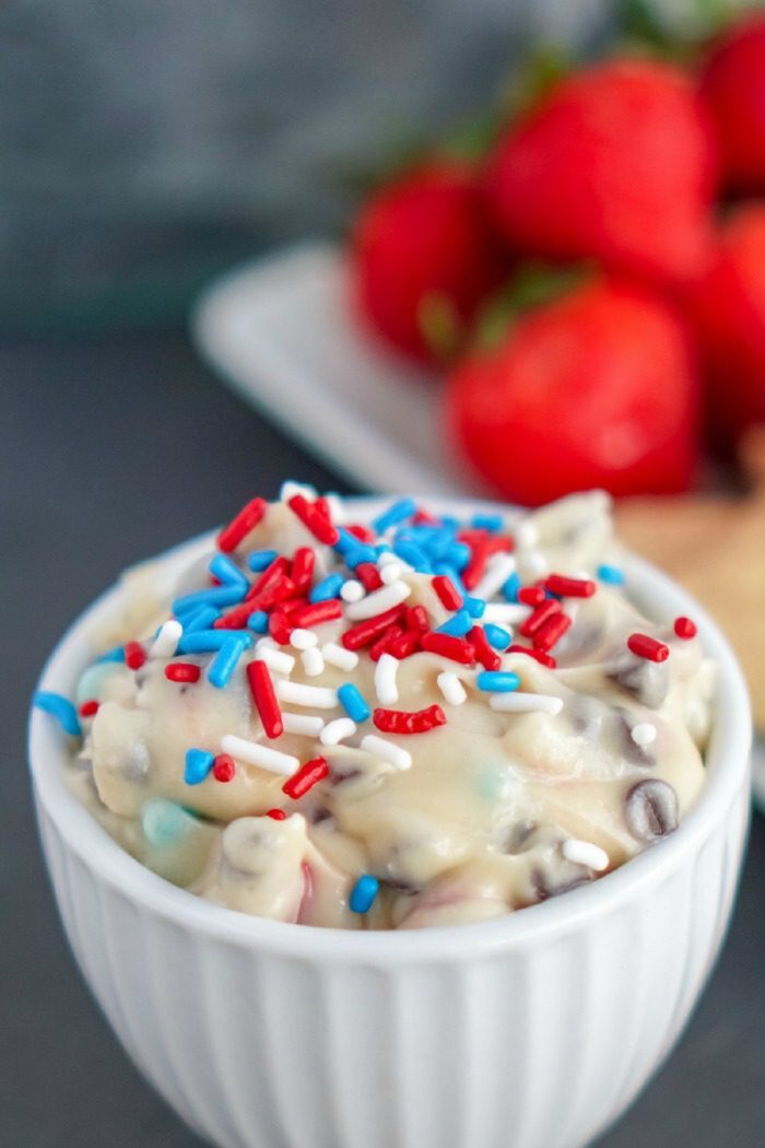 A bowl of cookie dough dip topped with red, white, and blue sprinkles sits in front of a plate of strawberries in the background.