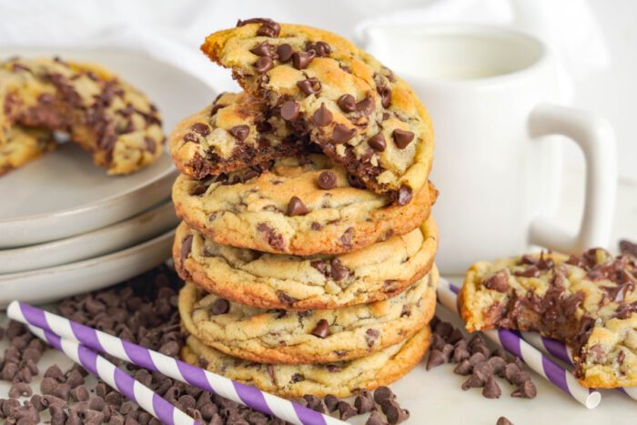 A stack of chocolate chip cookies on a table, with a mug of milk nearby. Purple and white striped straws and additional cookies surround the stack. One cookie has a bite taken out of it.