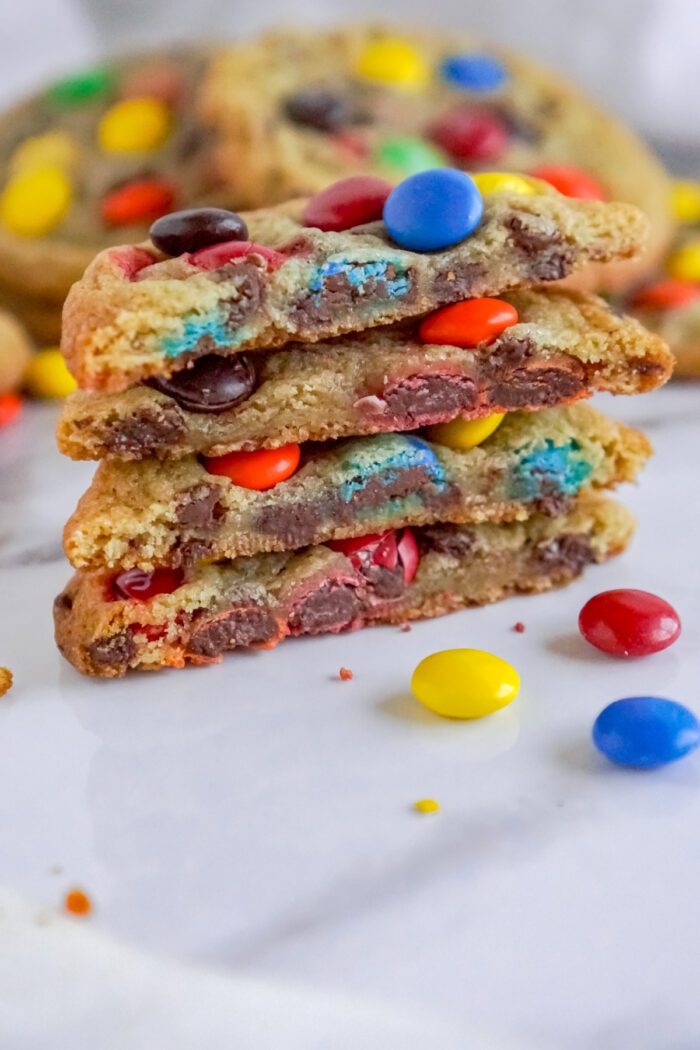 Stack of three sliced cookies with colorful chocolate candies on a white surface, with more cookies and candies in the blurred background.