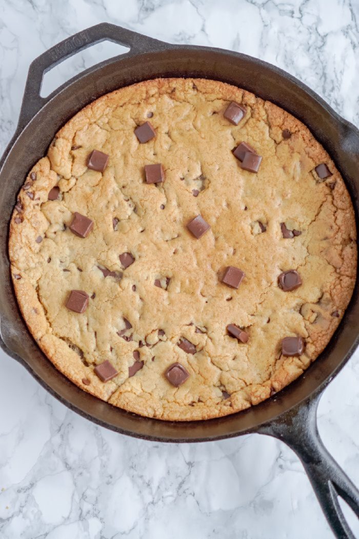 A large chocolate chip cookie baked in a cast iron skillet, topped with chocolate chunks, rests on a white marble countertop.