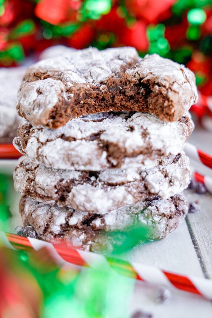 A stack of chocolate crinkle cookies with powdered sugar, one of which has a bite taken out of it, surrounded by festive red and green decorations.