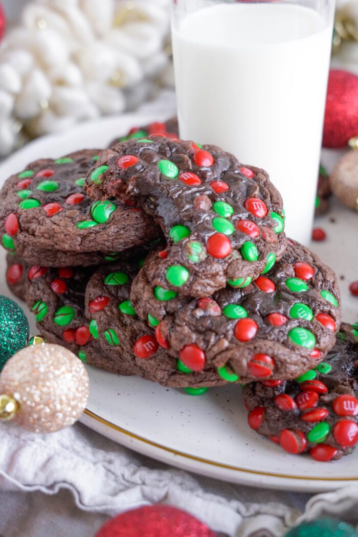 A plate of chocolate cookies with red and green candy pieces is placed beside a glass of milk. Holiday ornaments surround the plate.