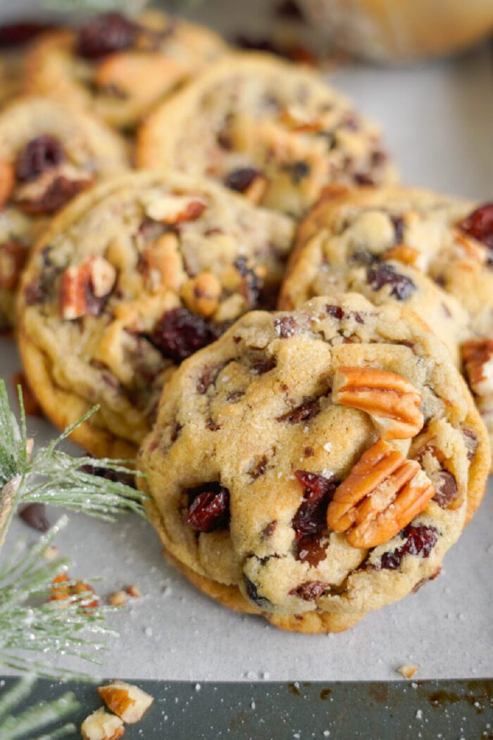 A close-up of several freshly baked cookies with pecans and dried cranberries on a surface. A small pine branch is visible to the left.