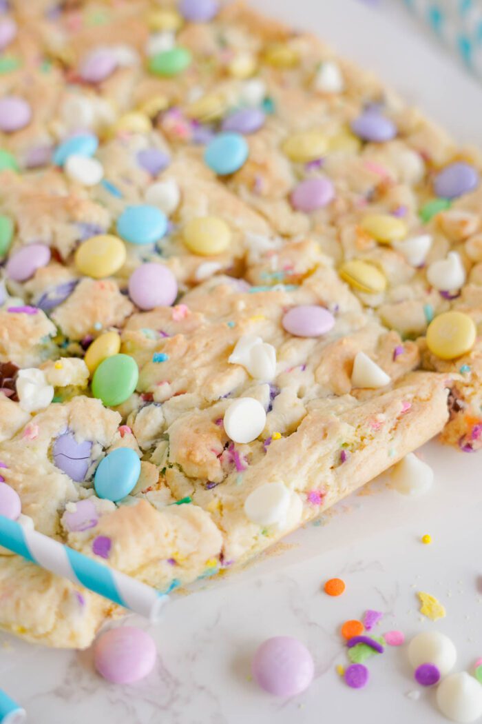 A close-up of a colorful cookie bar topped with white chocolate chips and pastel-colored candy pieces, placed on a white surface with scattered candy and two blue striped straws.