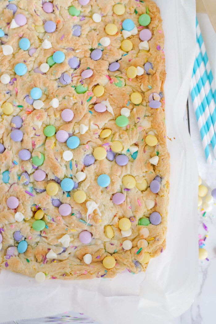 A large, baked cookie bar with colorful candy-coated chocolates and white chocolate chips on parchment paper. Striped straws and scattered candies are visible beside it, making these Easy Easter Cookie Bars with Cake Mix a delightful treat.