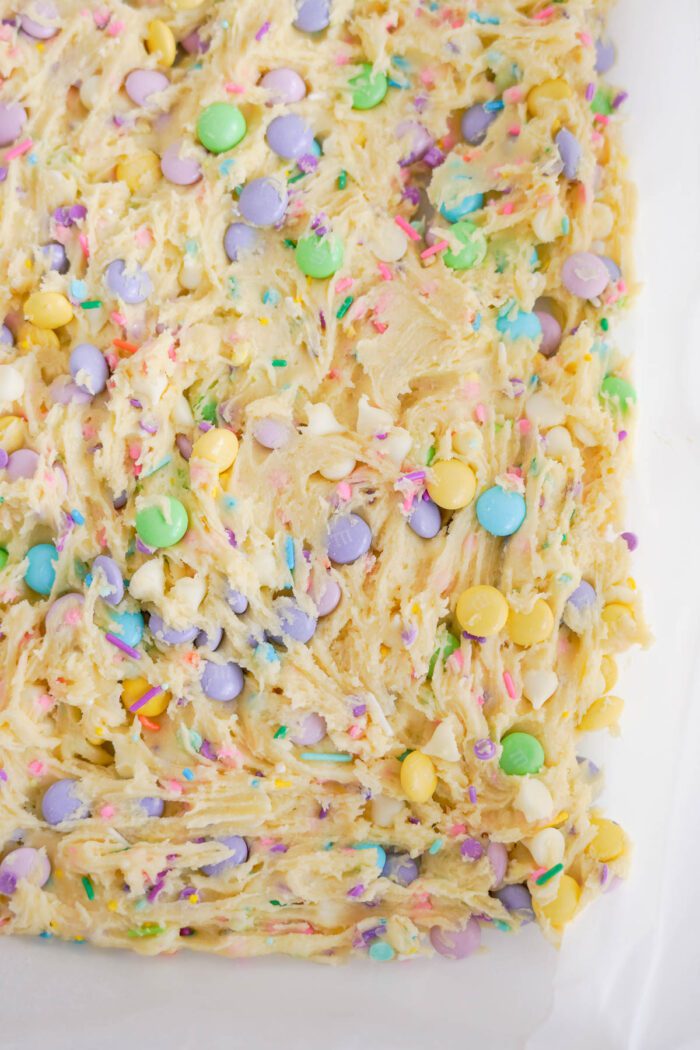 Close-up of unbaked cookie dough mixed with light purple, green, yellow, and white candy-coated chocolates and rainbow sprinkles on a parchment-lined baking tray.