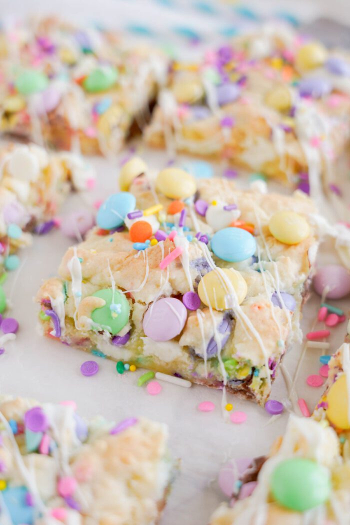 A close-up of Easter cookie bars topped with pastel candies and sprinkles, drizzled with white icing, on a white surface.