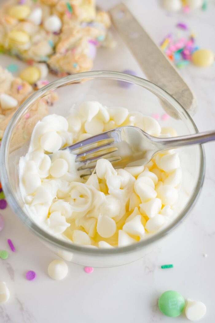 A clear glass bowl containing white chocolate chips and a metal fork. Scattered around are various colorful sprinkles and confectioneries.