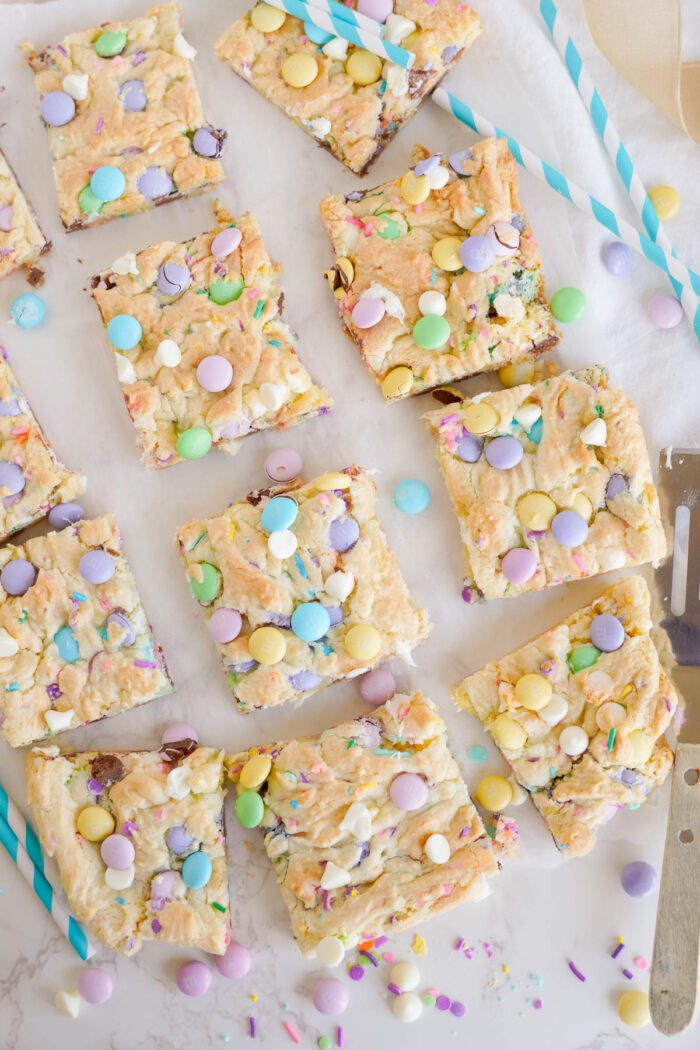 Squares of bar cookies topped with colorful candies are arranged on a white surface. Scattered around are more candies and striped blue straws. A metal spatula is located to the right.