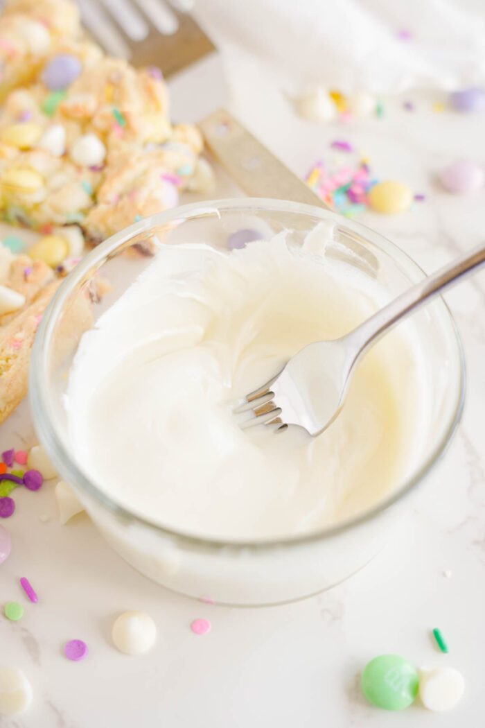 A glass bowl filled with white frosting and a fork, surrounded by colorful sprinkles and candies on a white surface.