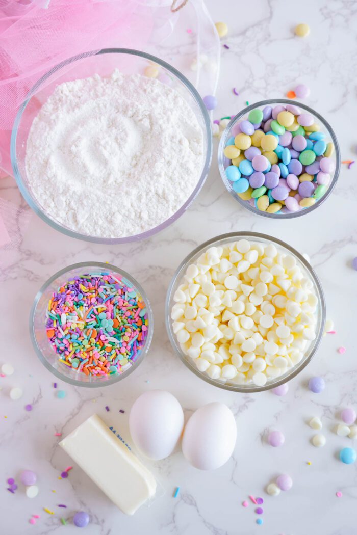 Ingredients for baking laid out on a marble surface: powdered sugar, M&Ms, Easter sprinkles, white chocolate chips, two eggs, and a stick of butter.