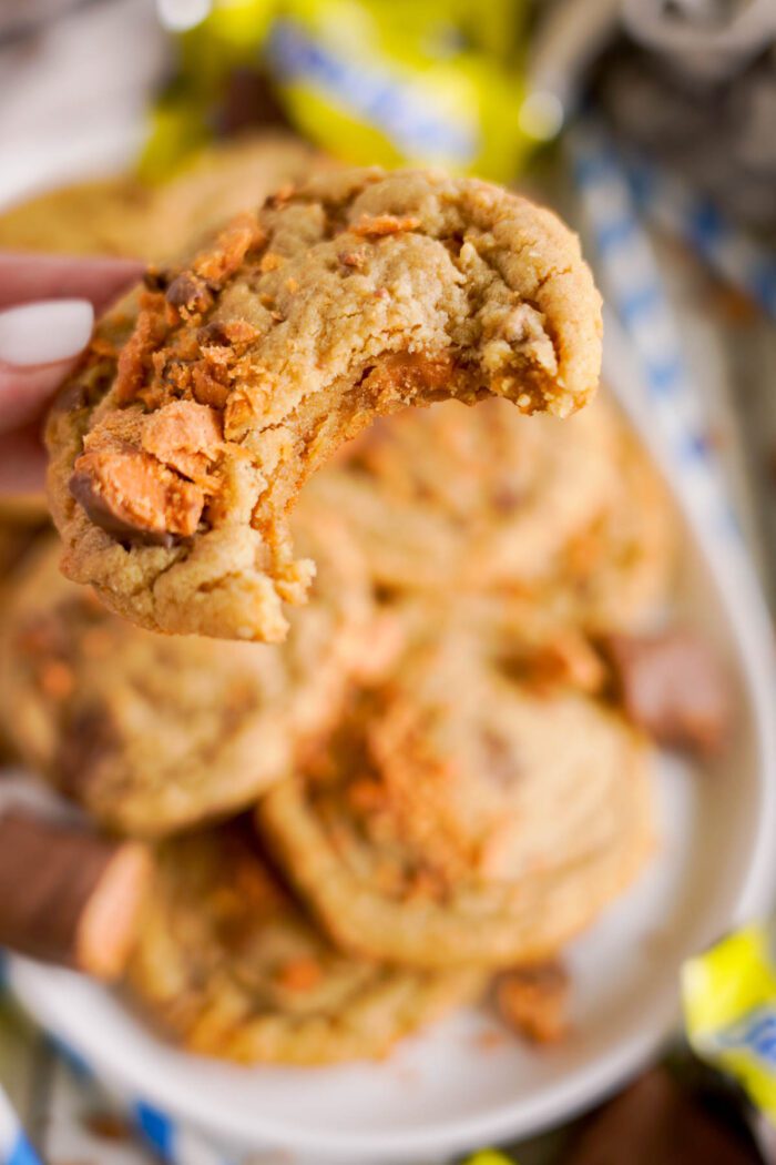 A close-up of a hand holding a bitten cookie with visible chunks of butterfinger candy. More cookies and candy are on a white plate in the background.