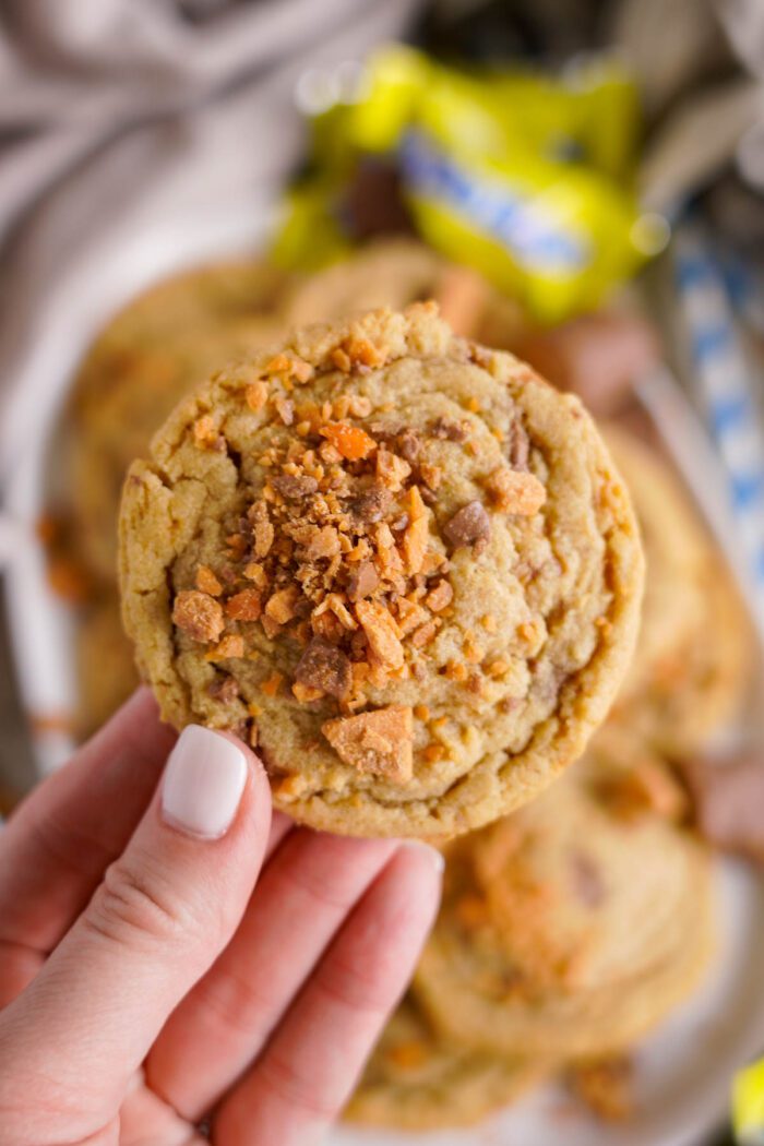 A hand holding a cookie topped with crushed butterfinger bits. Other cookies are blurred in the background.