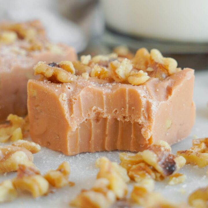 Close-up of a piece of butterscotch fudge topped with walnuts and a bite taken out. More walnuts are scattered around on a white surface.