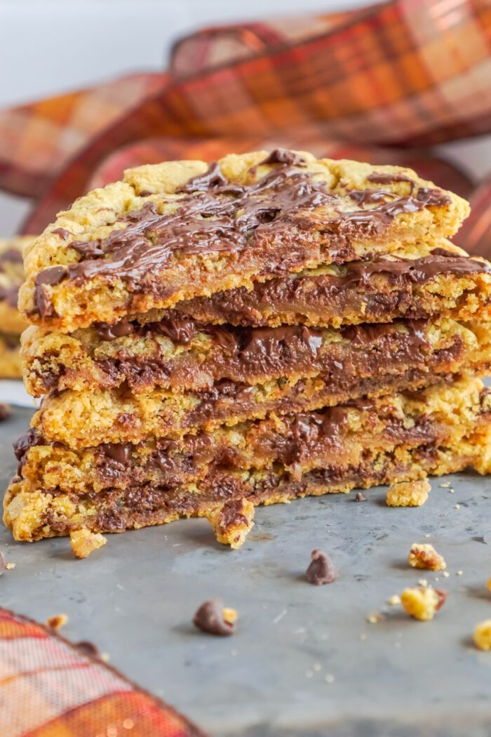 A stack of chocolate chip cookies with layers of melted chocolate in between, placed on a gray surface with a plaid ribbon in the background.