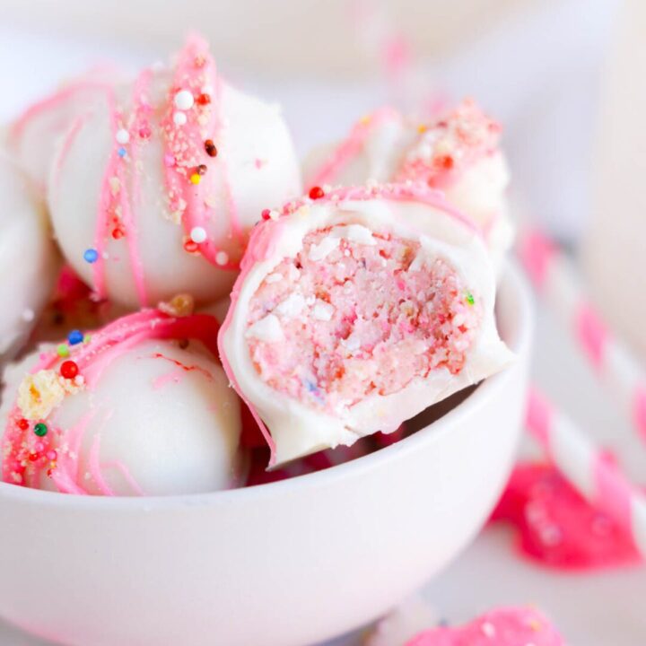 A white bowl contains white chocolate-covered cake balls with pink drizzle and colorful sprinkles. One cake ball is cut in half, revealing a pink, crumbly inside.