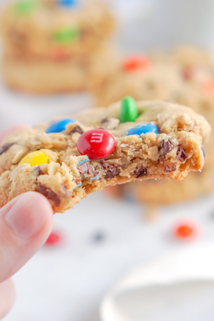 A hand holds a bitten cookie decorated with colorful chocolate candies. Other cookies and candies are blurred in the background.