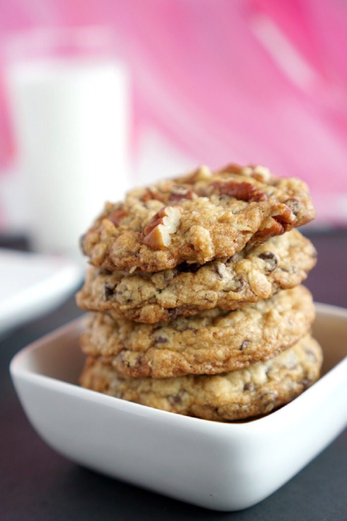 A stack of four cookies with nuts in a white square bowl, set on a dark surface. In the blurry background, a glass of milk is visible against a pink patterned backdrop.