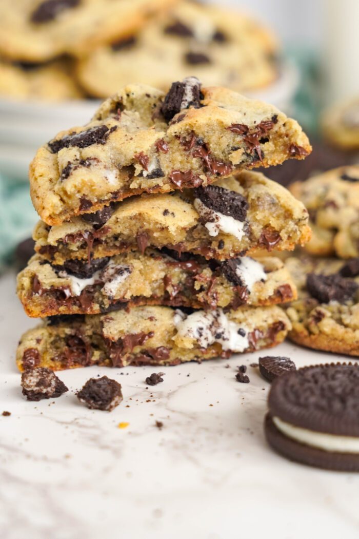 A close-up of chocolate chip cookies with Oreo pieces, stacked and broken in half, revealing gooey chocolate and marshmallow filling. More cookies and Oreo biscuits are in the background.